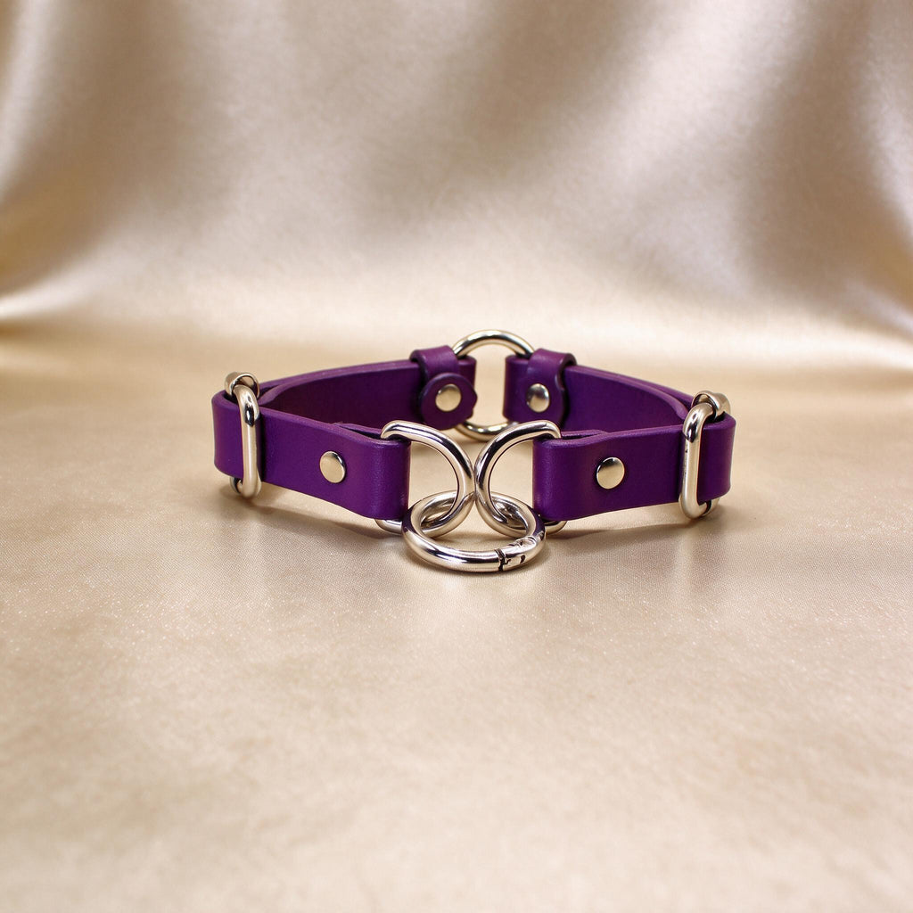 A purple leather choker necklace with silver metal rings and buckles is displayed on a beige background.