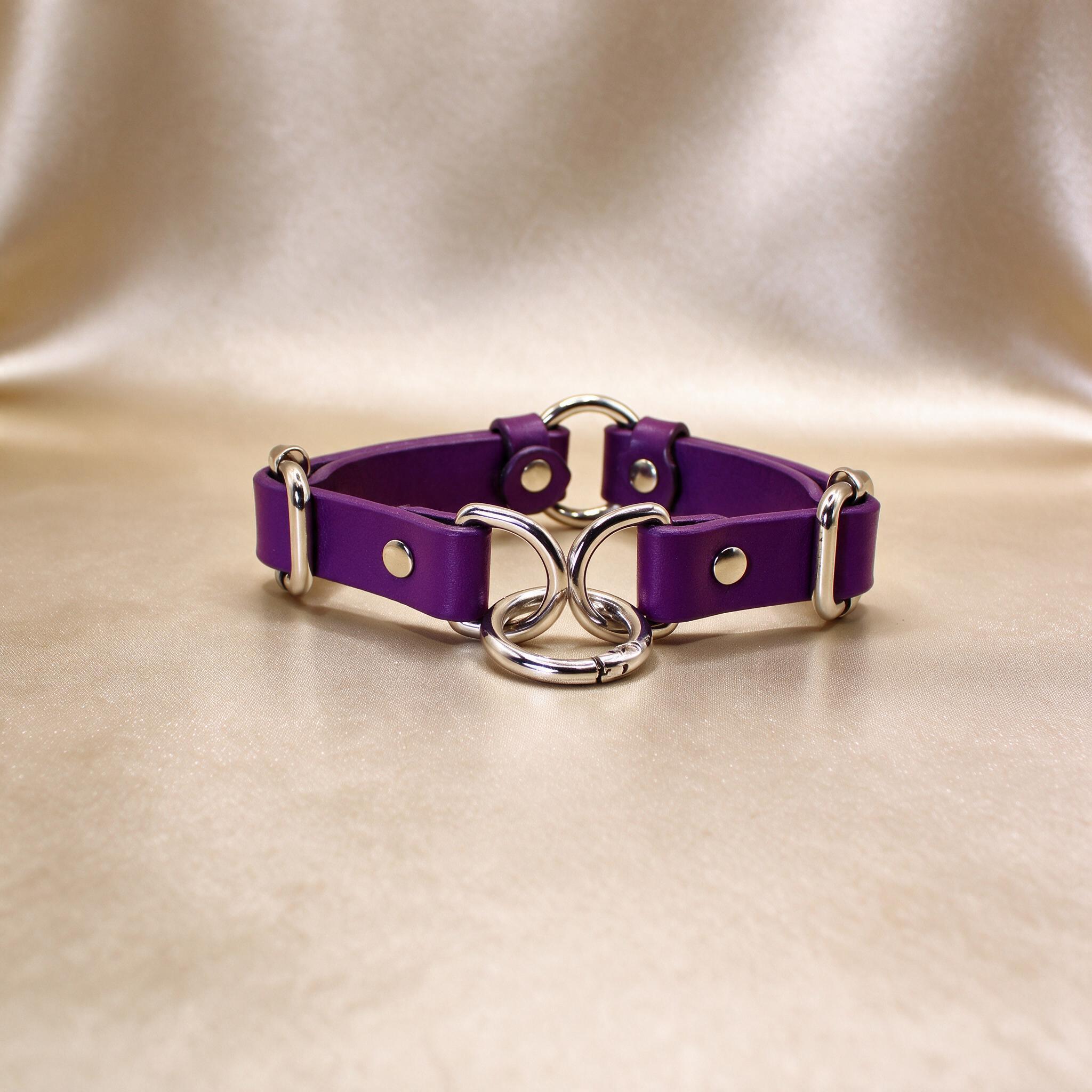 A purple leather choker necklace with silver metal rings and buckles is displayed on a beige background.