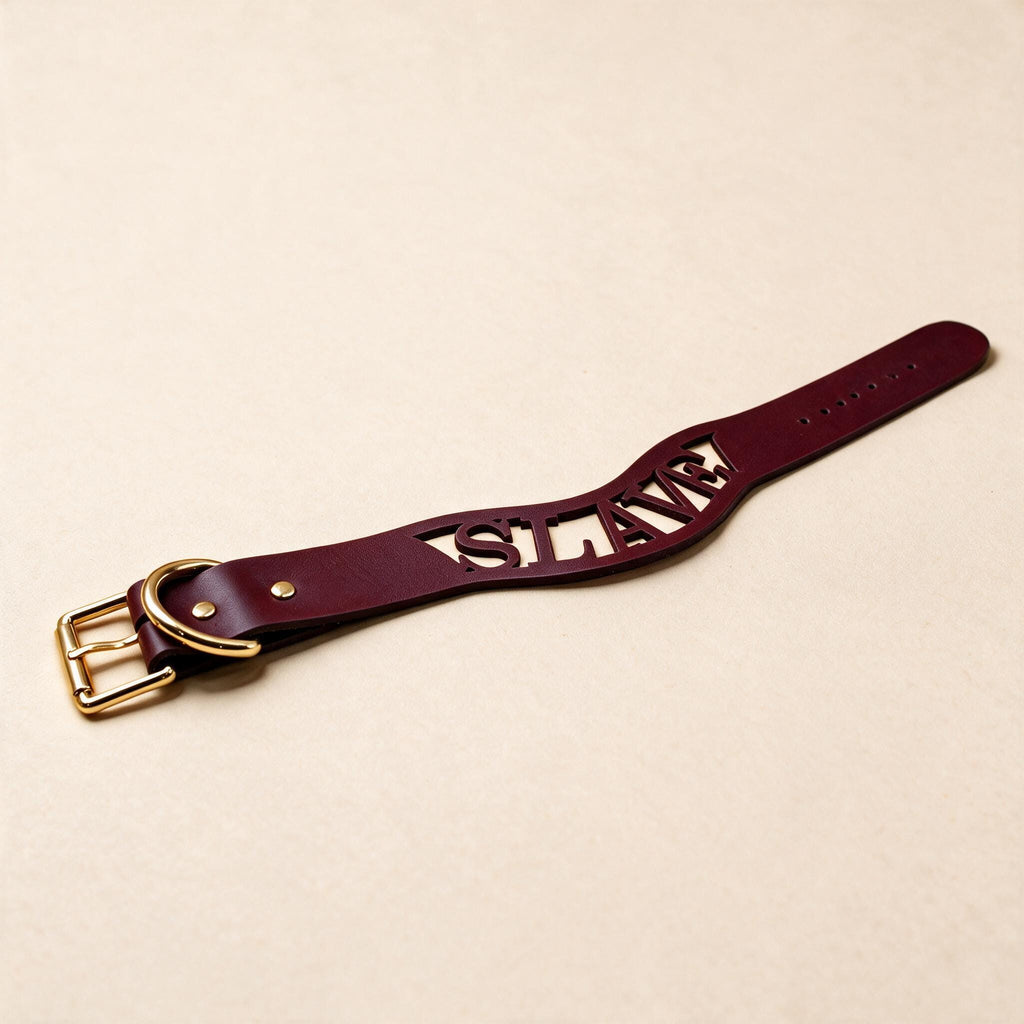 A burgundy leather belt with the word "SLAVE" carved into it, lying on a beige surface.