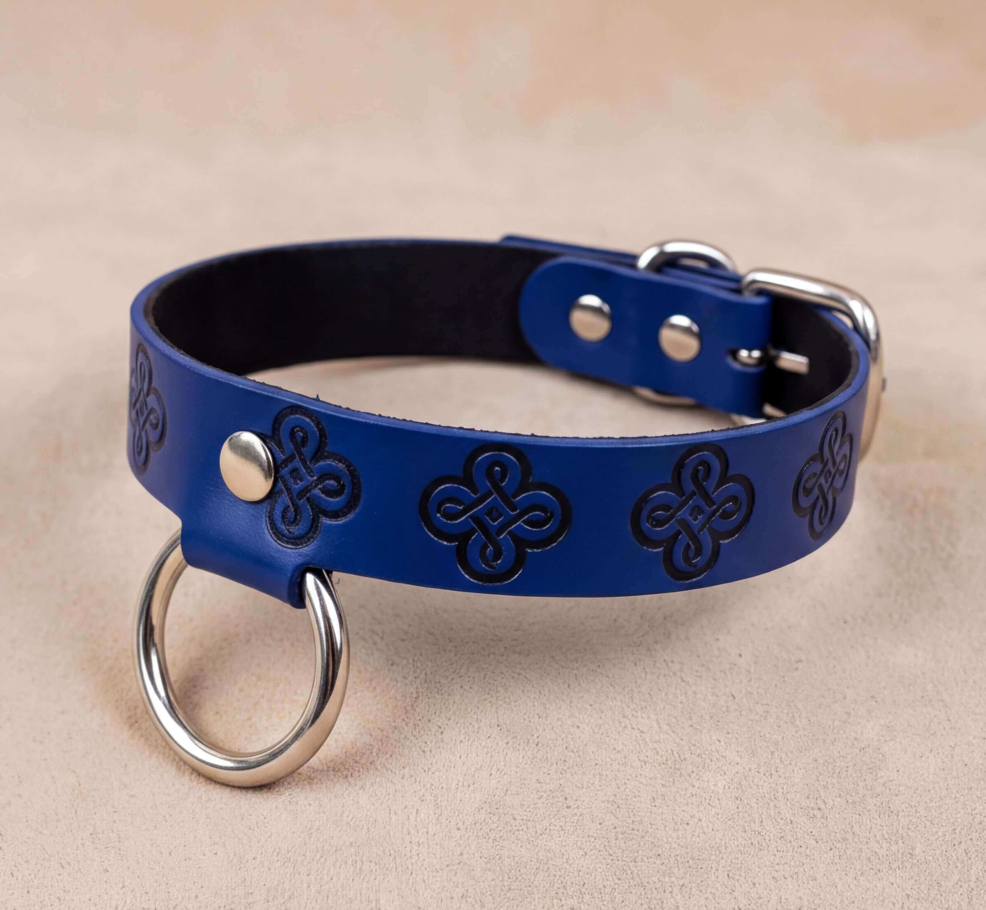 A blue leather dog collar with metal rings and a buckle, placed on a beige surface.