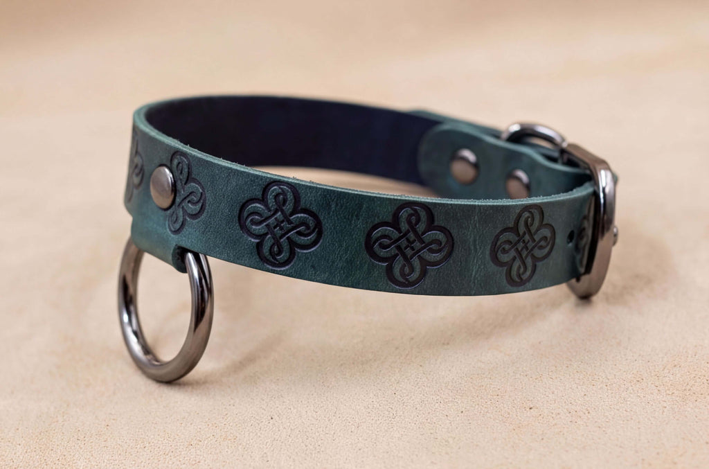 A green leather dog collar with metal rings and a buckle, resting on a beige surface.