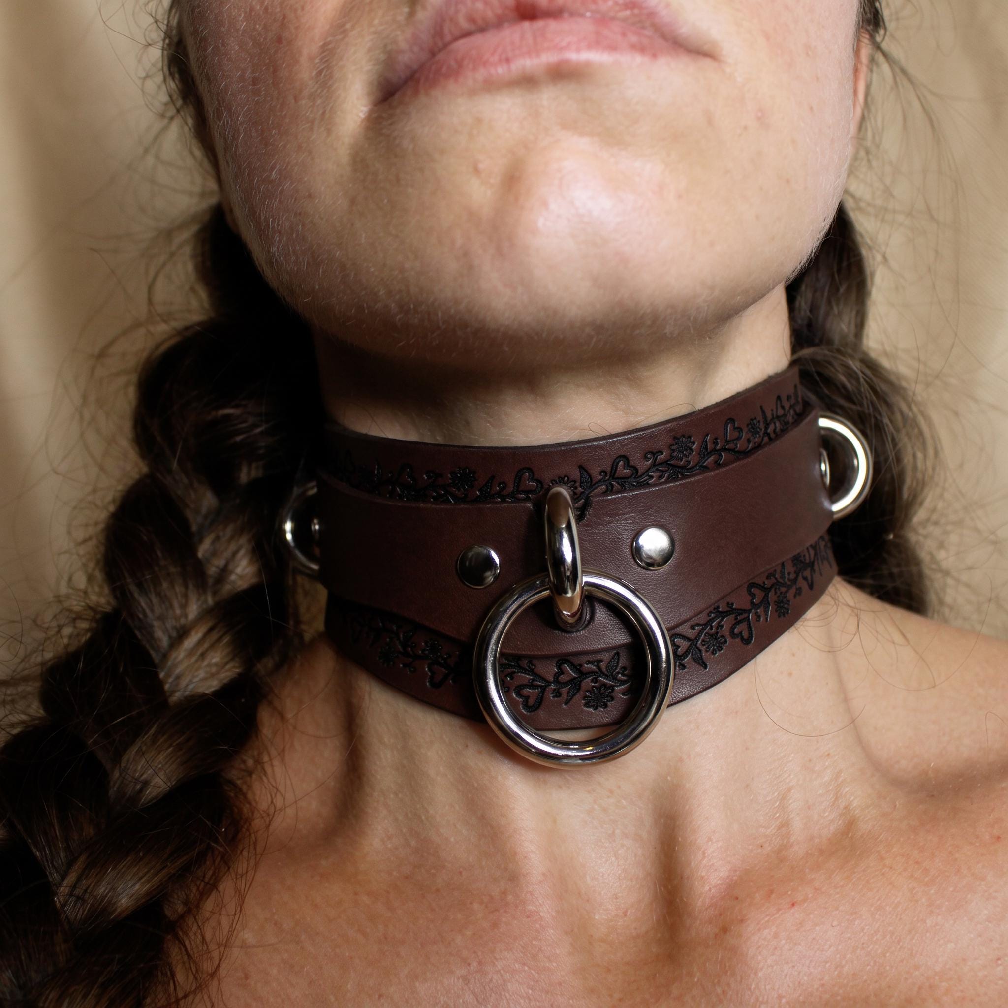 A close-up of a person's neck wearing a brown leather choker necklace with metal rings and a circular clasp.
