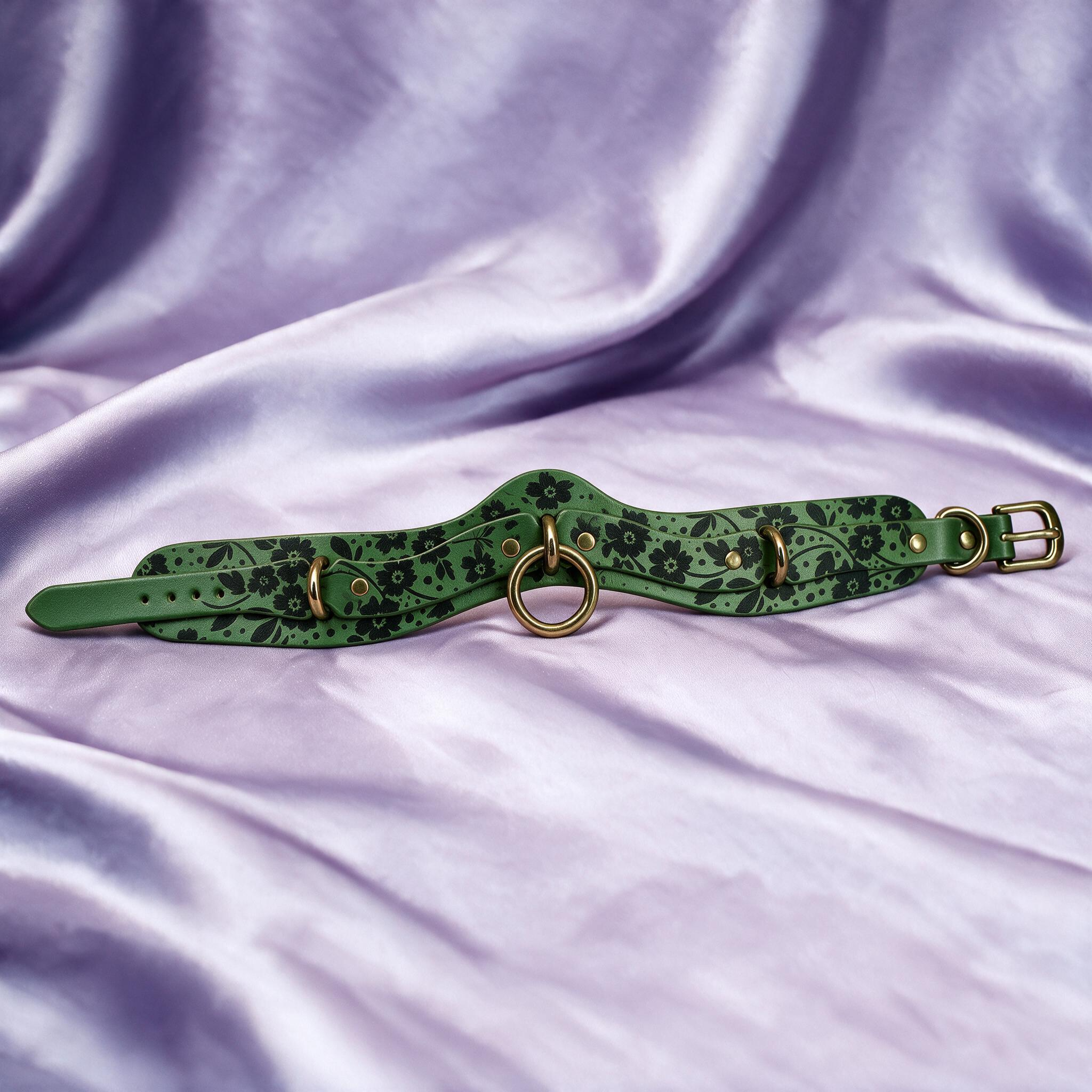 A green leather choker necklace with floral patterns and metal rings is displayed on a purple fabric background.
