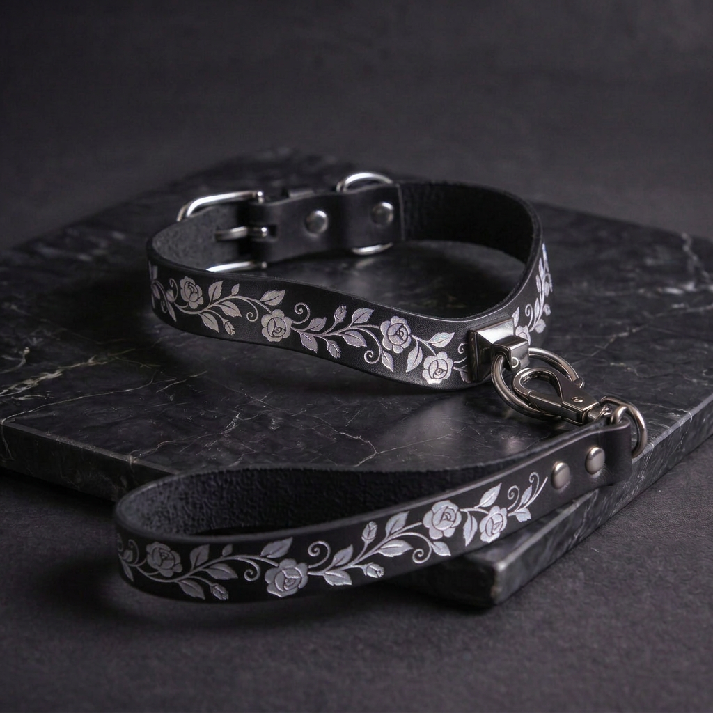 A black leather collar with a floral pattern and a matching leash attached to it, resting on a dark surface.