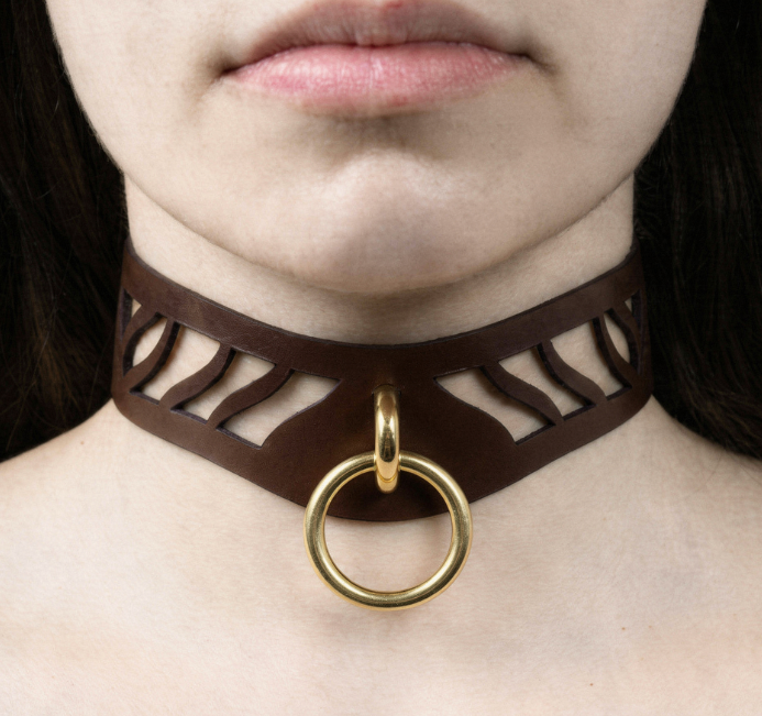 A close-up of a person's neck and chest area, featuring a brown leather choker necklace with a gold ring and a cutout design.