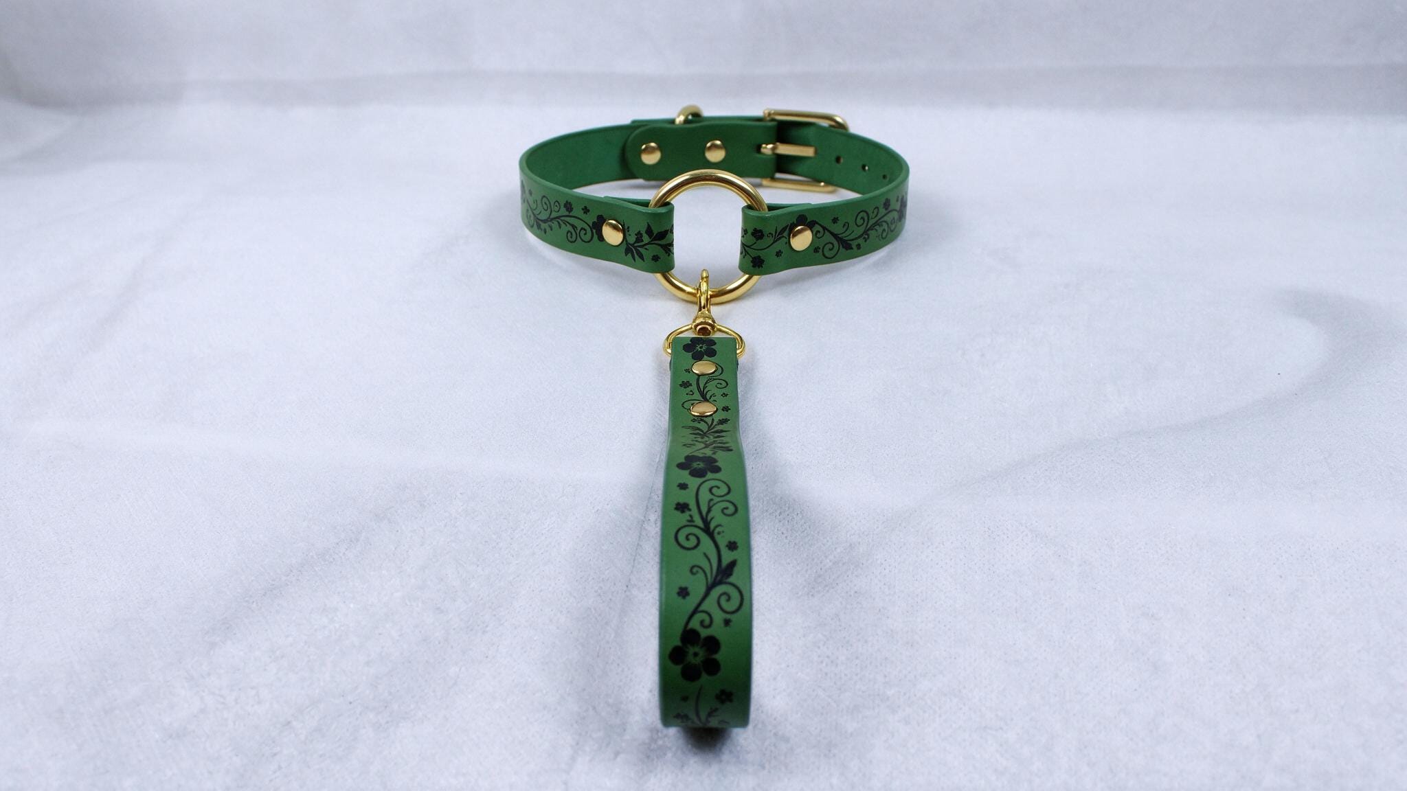 A green collar with a floral pattern and a gold-colored buckle is displayed on a white surface.