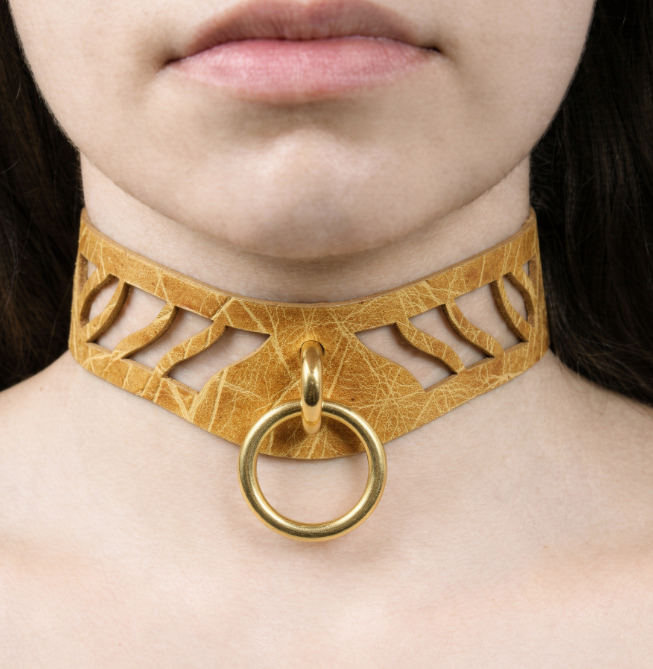 A close-up of a woman's neck and chest area wearing a gold-colored choker necklace with a circular ring.