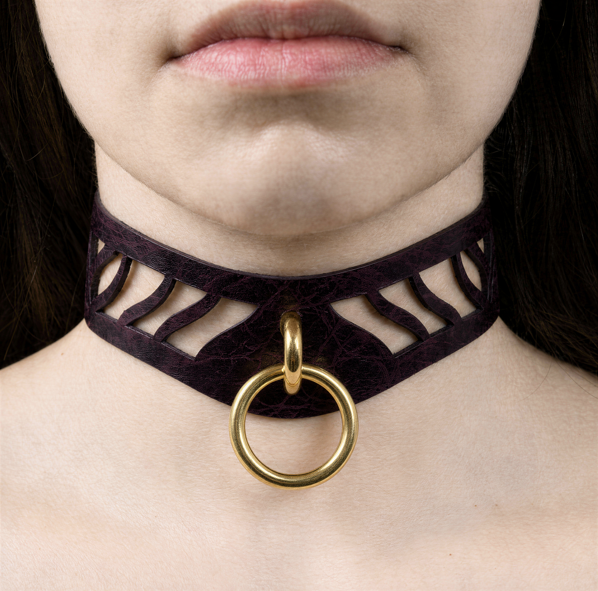 A close-up of a person's neck and chest area, featuring a dark purple choker necklace with a gold ring and a cutout design.