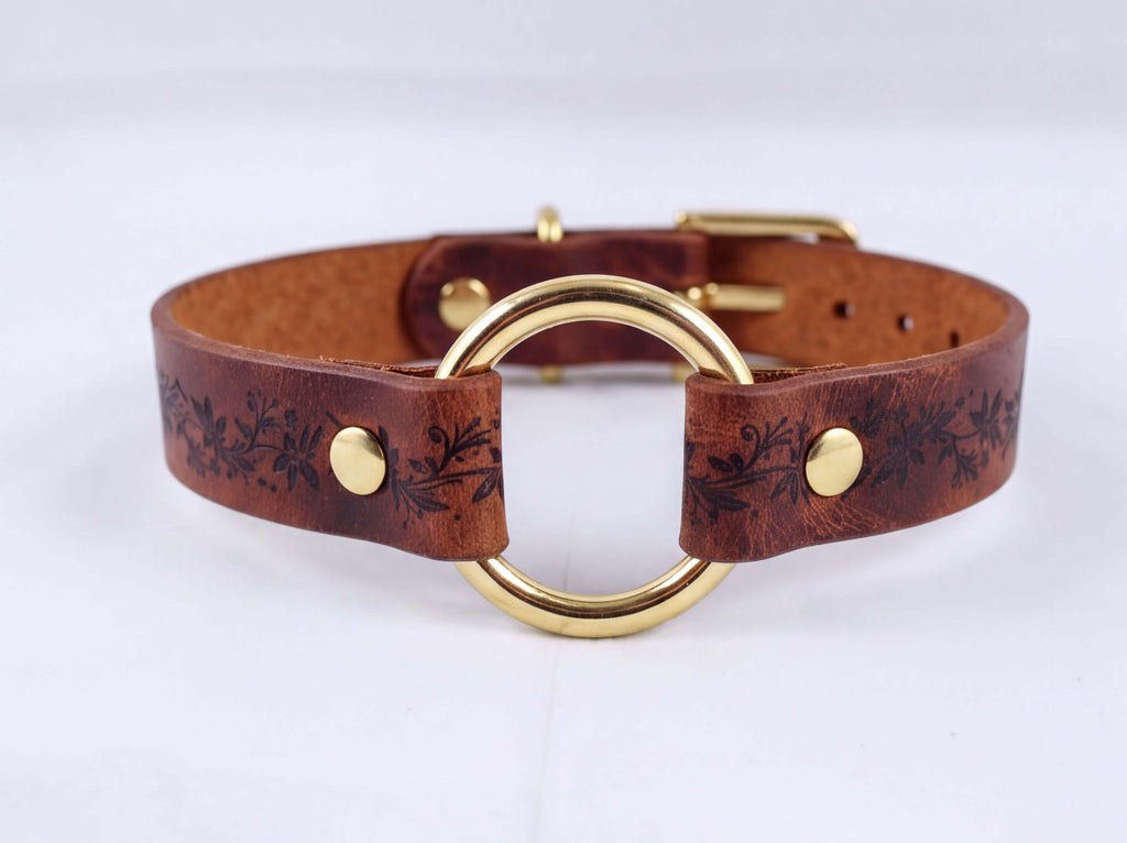 A brown leather choker collar with a gold-colored metal ring and decorative floral patterns.