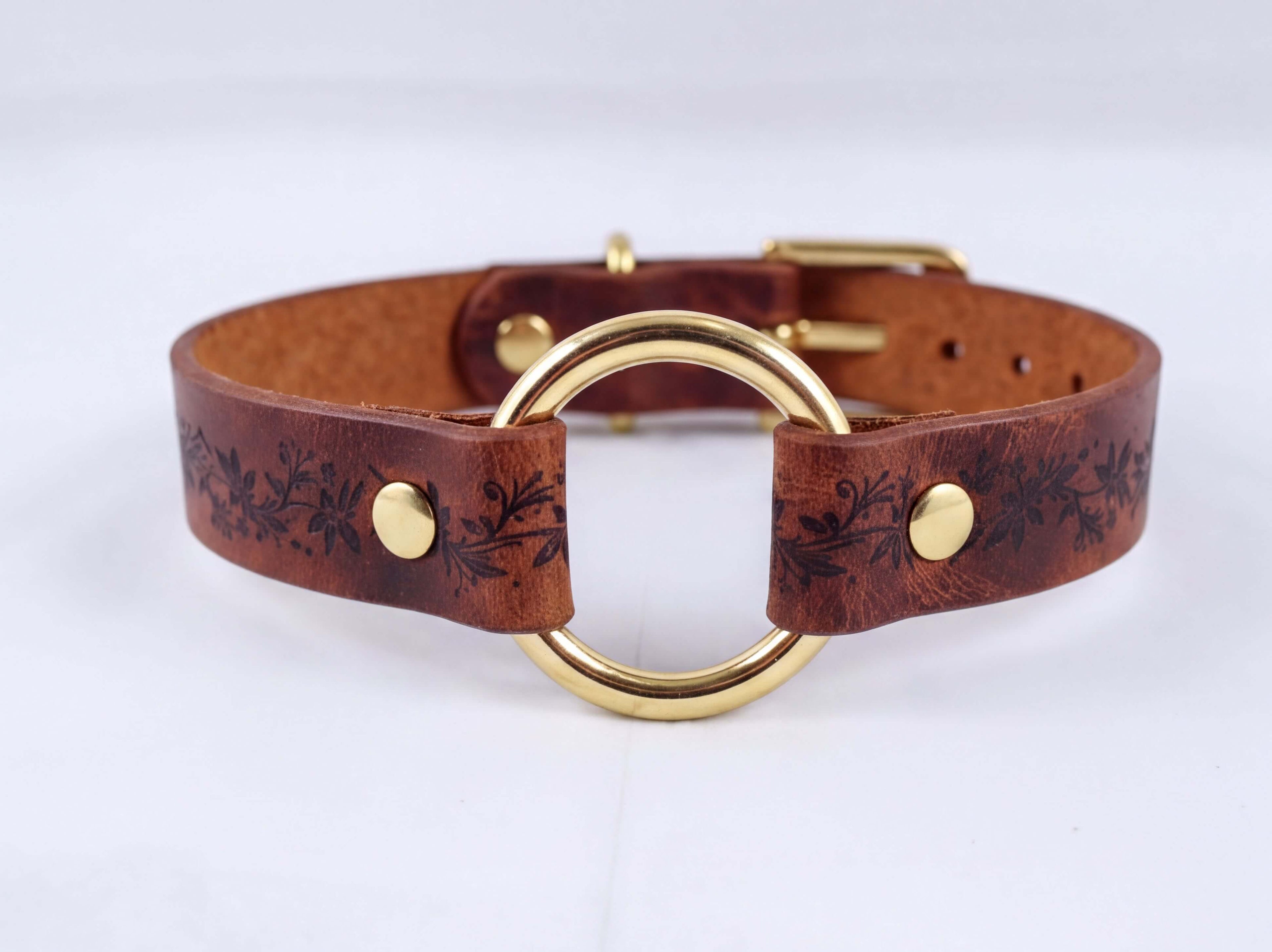 A brown leather choker collar with a gold-colored metal ring and decorative floral patterns.
