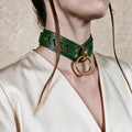 A close-up of a person wearing a green leather choker necklace with a gold ring, against a plain background.