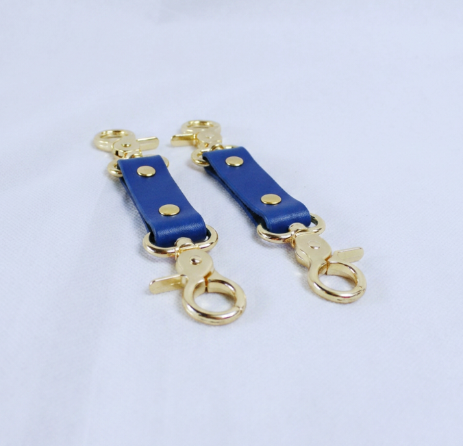 A pair of blue leather straps with gold metal rings and clasps, placed on a white surface.