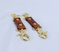 A pair of gold-colored leather keychains with metal clasps and loops, resting on a white surface.