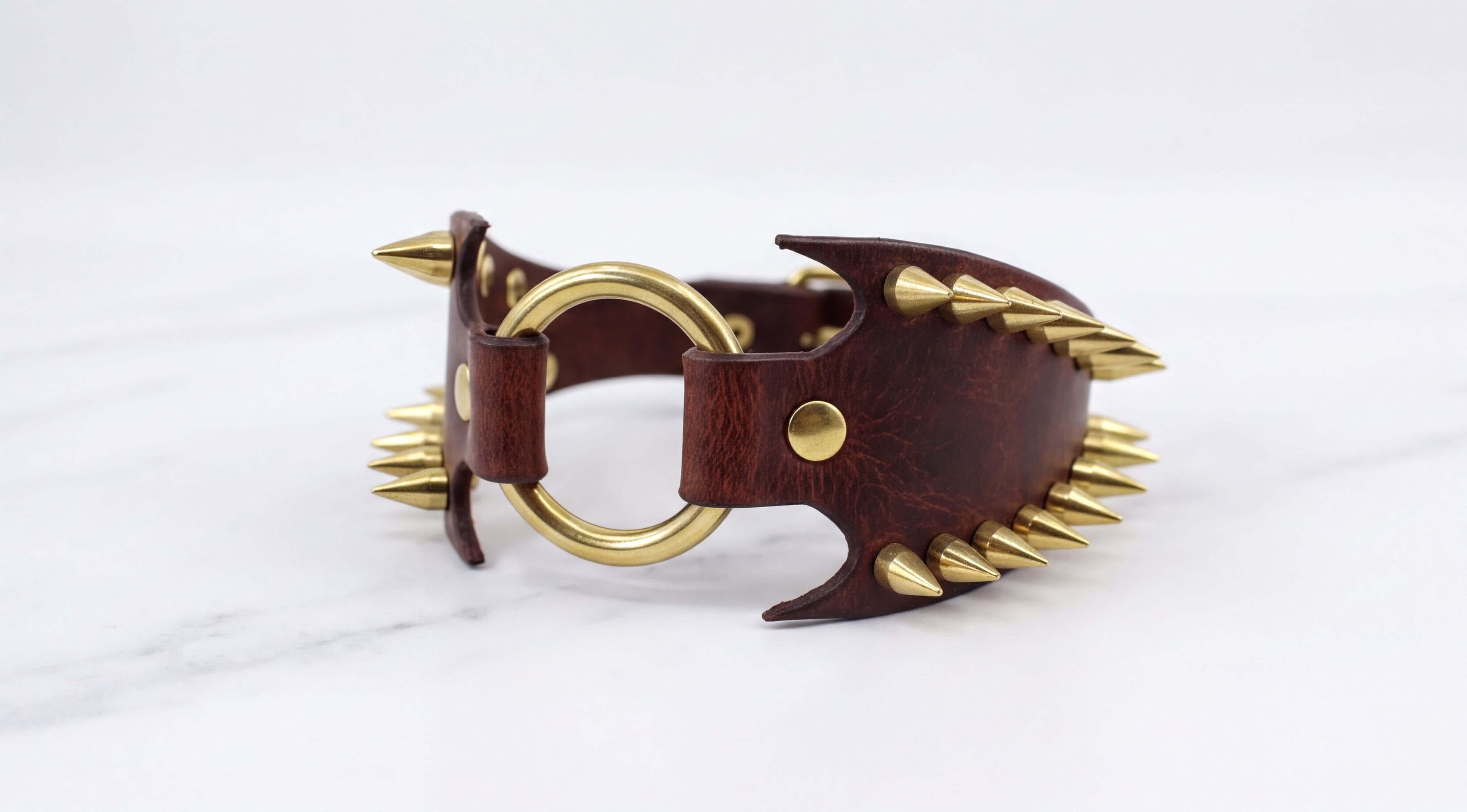 A brown leather bracelet with gold spikes protruding from it, resting on a white marble surface.