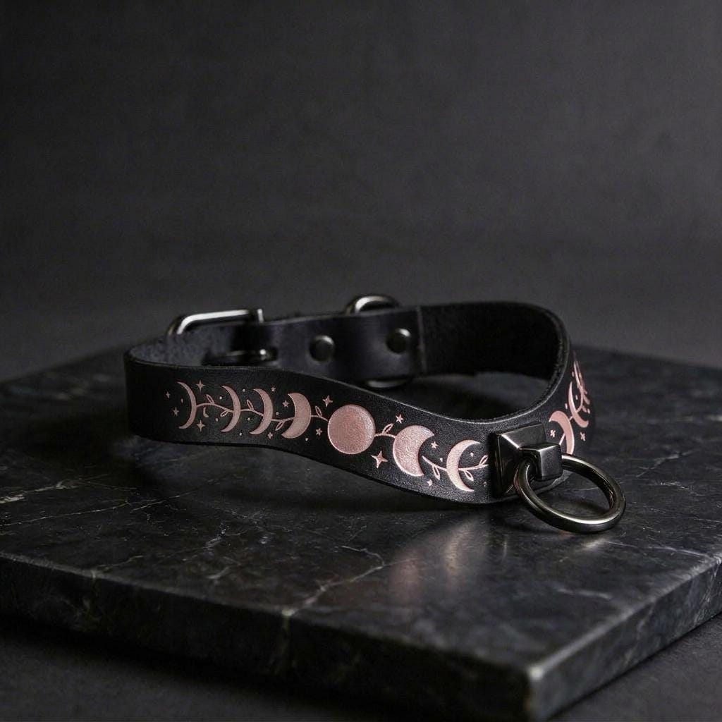 A black leather collar with a metallic ring and a decorative pattern of moon phases and stars on it, resting on a dark surface.