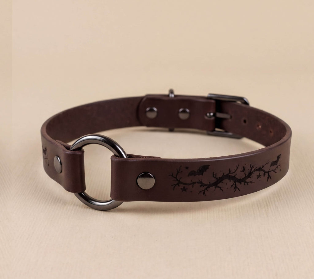 A brown leather collar with a metal ring and a design featuring bats and branches.