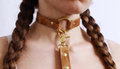 A close-up of a person's neck and chest area, featuring a leather choker collar with a gold-colored metal clasp and a leather leash attached to it.