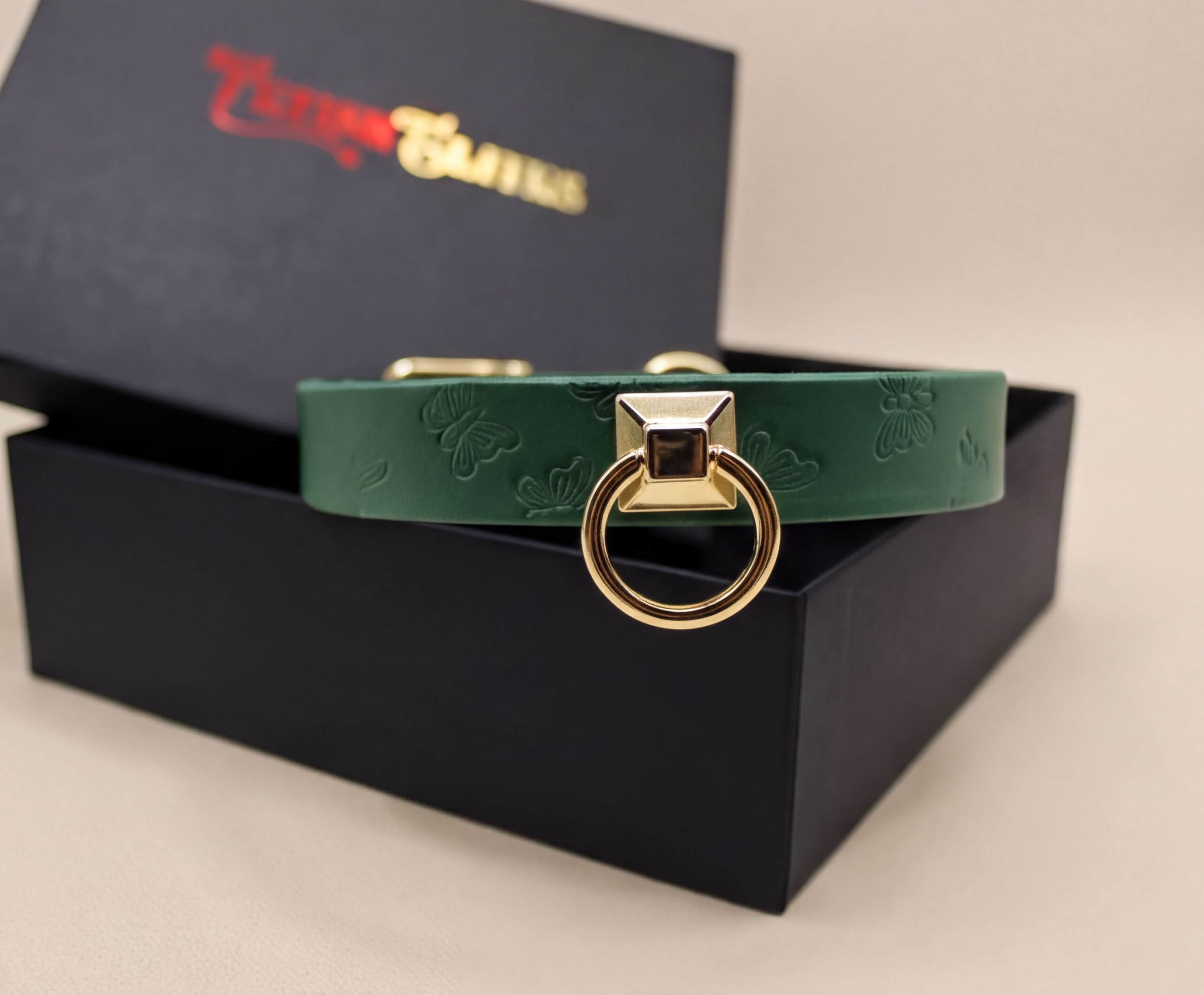 A green leather collar with a gold-colored metal ring and a black box with a red logo on it.