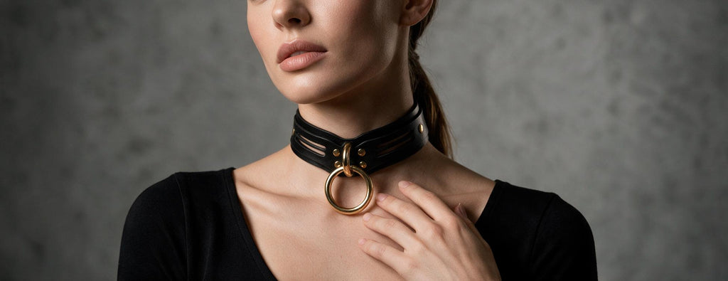 A woman wearing a black choker necklace with a gold ring on it, posing against a gray background.