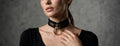 A woman wearing a black choker necklace with a gold ring on it, posing against a gray background.