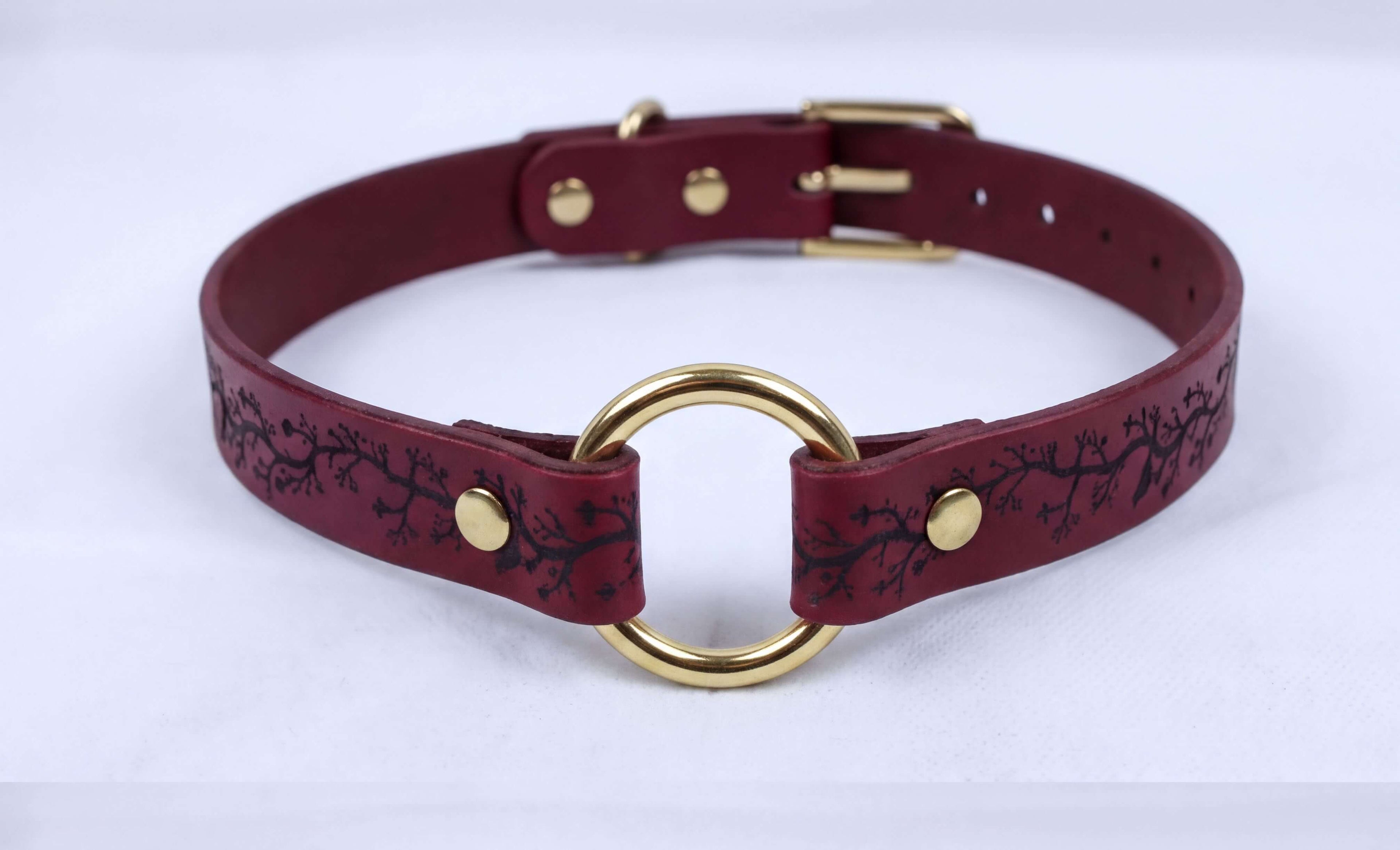 A burgundy leather collar with a gold-colored ring and a black tree-like design on the strap.