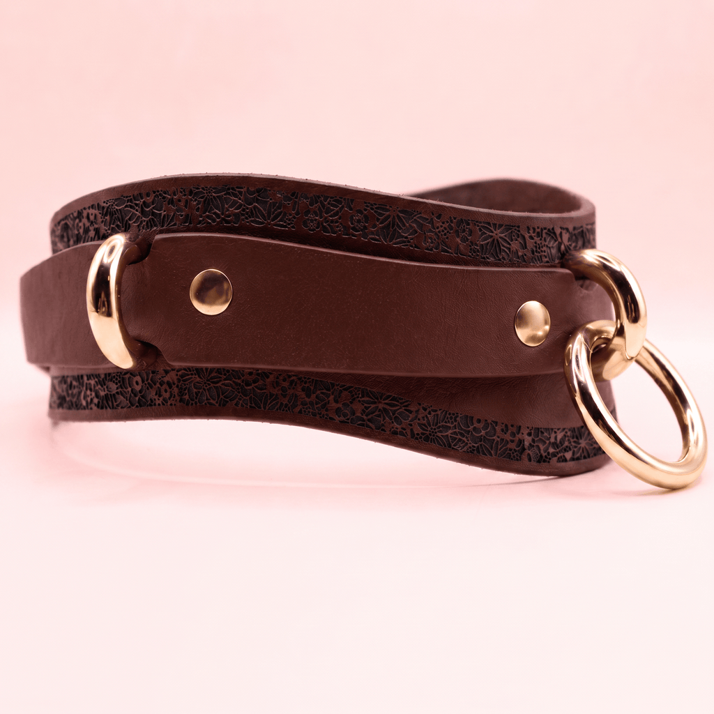 A brown leather belt with a gold-colored buckle and two round rings on the ends, set against a pink background.