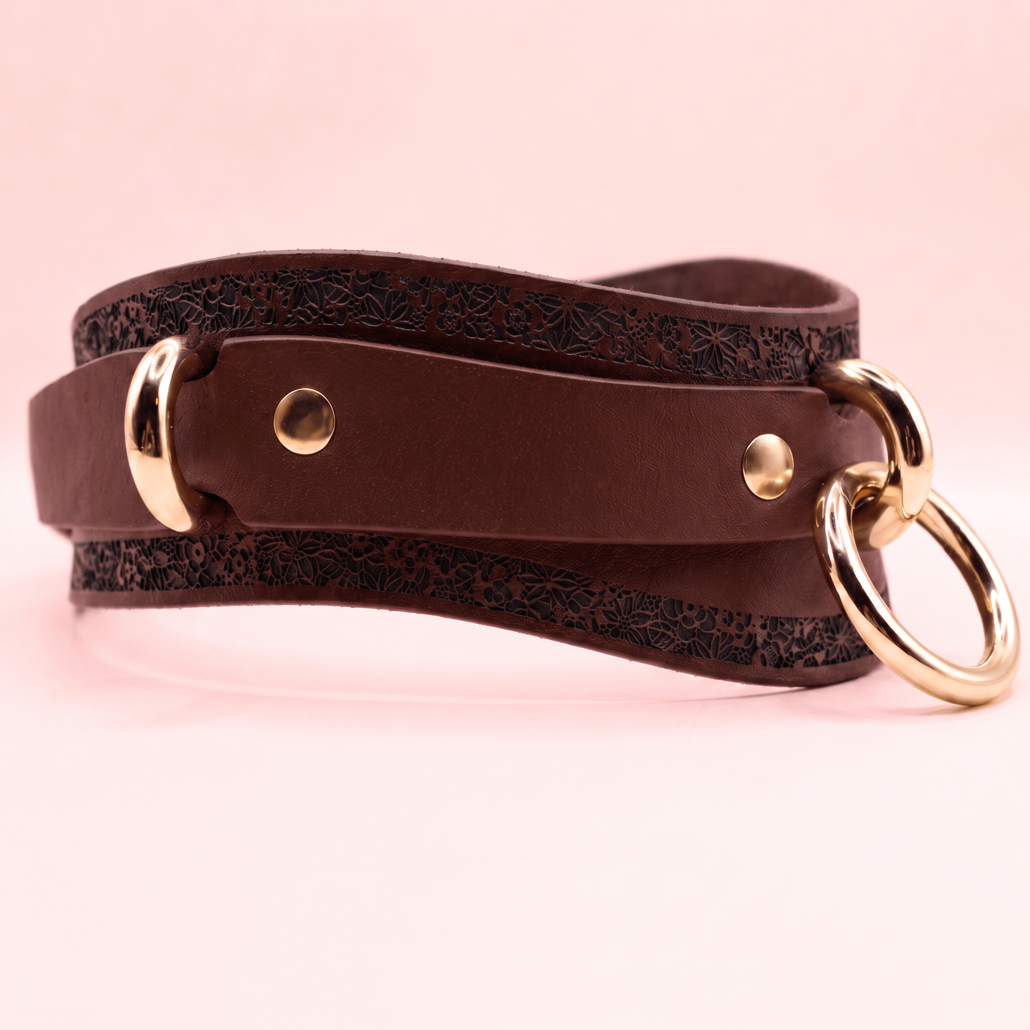 A brown leather belt with a gold-colored buckle and two round rings on the ends, set against a pink background.