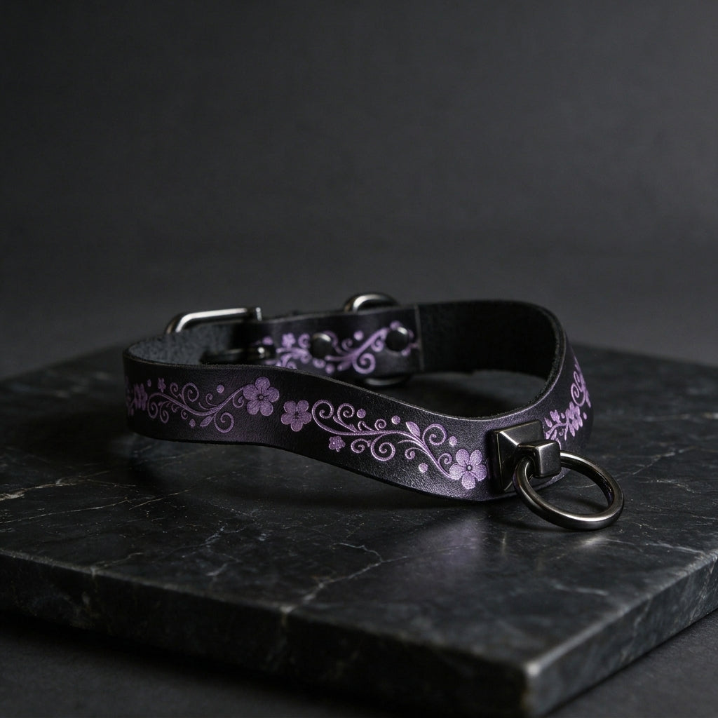 A black leather collar with a floral pattern and a metal ring on it, resting on a dark surface.