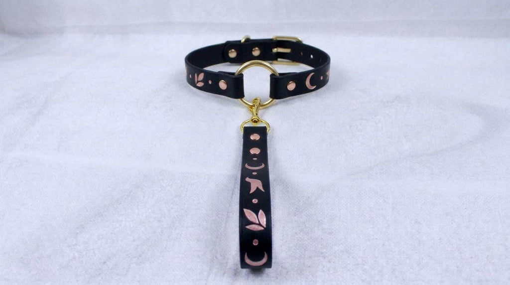 A black leather collar with a gold-colored metal clasp and a decorative metal handle attached to it, resting on a white surface.
