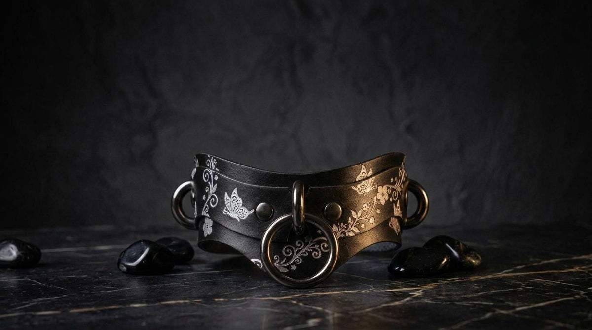 A black and gold leather collar with decorative elements, including a circular metal ring, is displayed on a dark surface with small black stones.