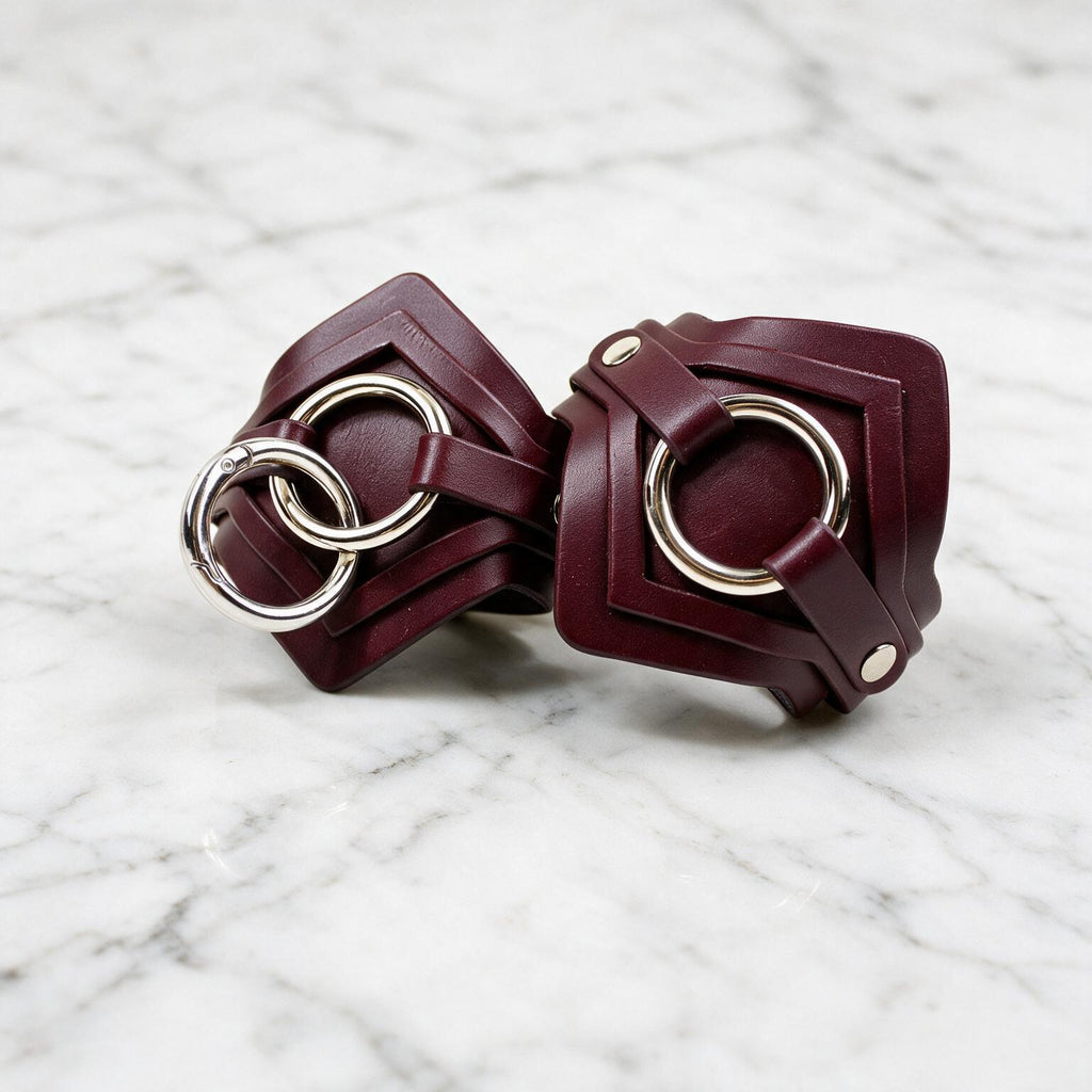 A pair of burgundy leather wristbands with metal rings and buckles are displayed on a marble surface.
