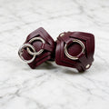 A pair of burgundy leather wristbands with metal rings and buckles are displayed on a marble surface.