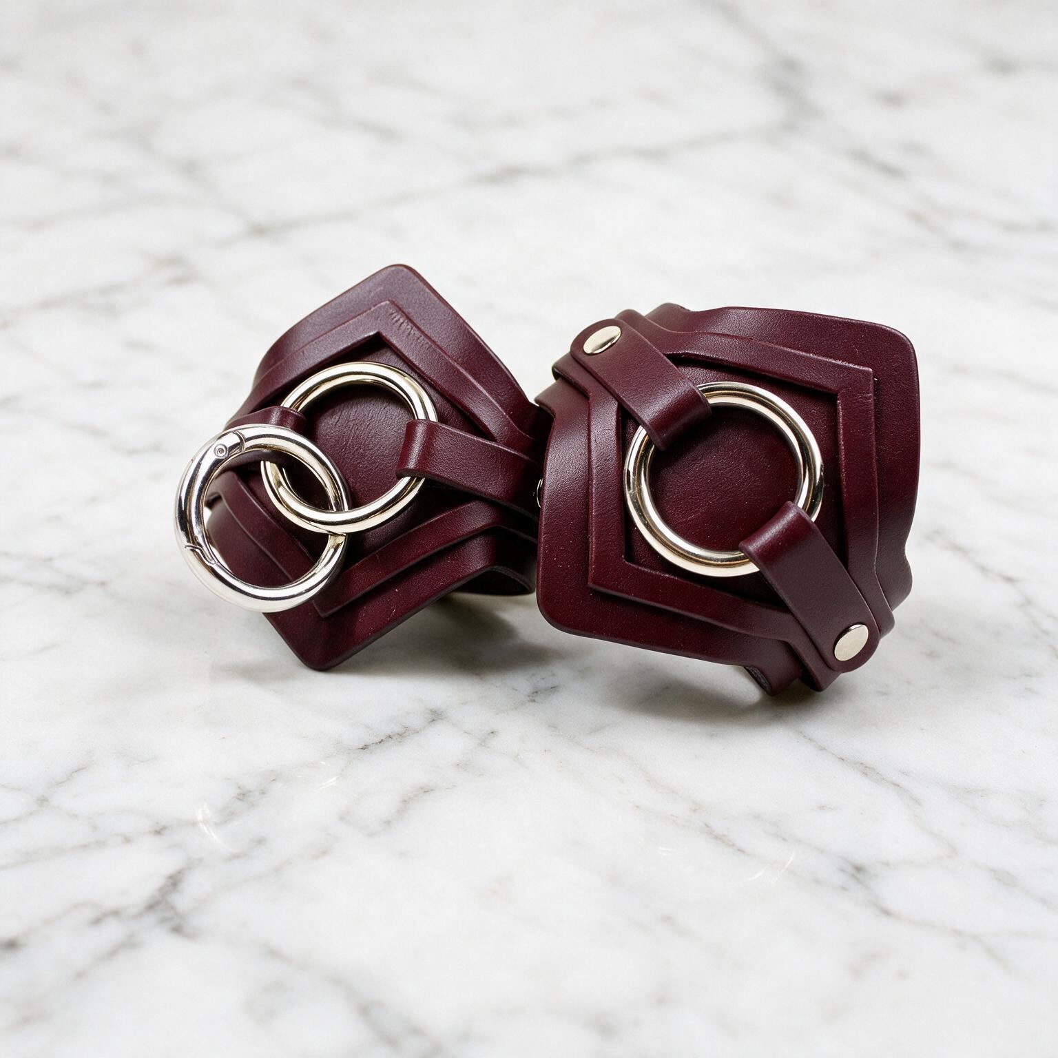 A pair of burgundy leather wristbands with metal rings and buckles are displayed on a marble surface.