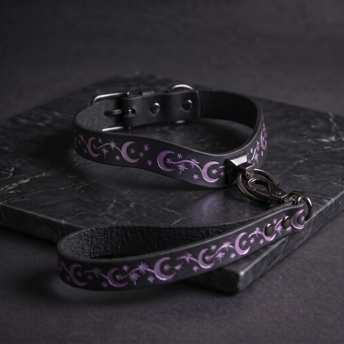 A black leather dog collar with a purple star and crescent design, and a matching leash, resting on a dark surface.