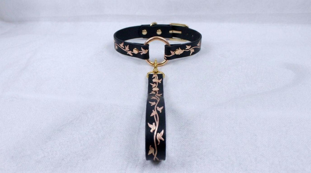 A black collar with a gold-colored metal clasp and a decorative floral pattern on the strap.