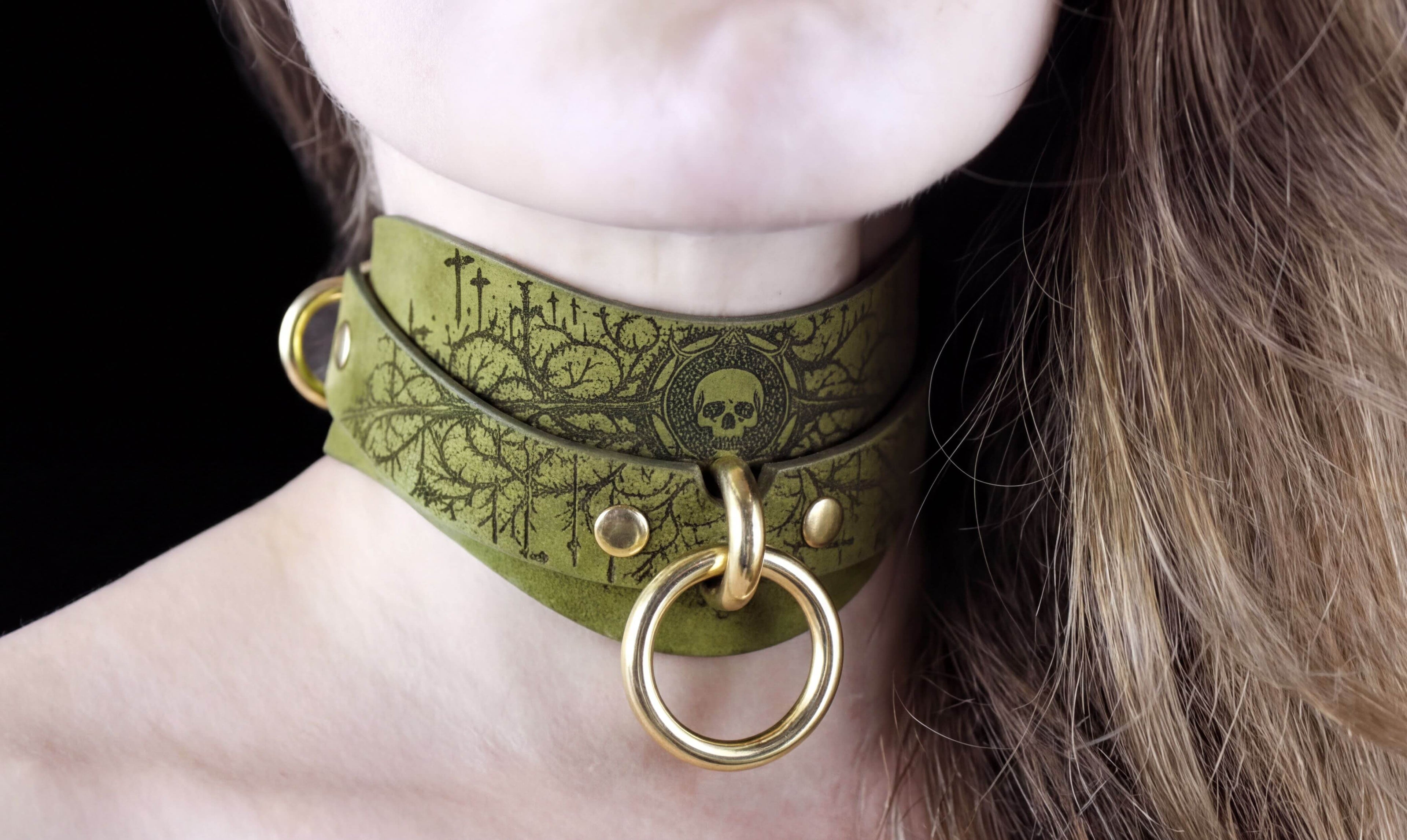 A close-up of a woman's neck and chin wearing a green choker necklace with a skull design and gold rings.