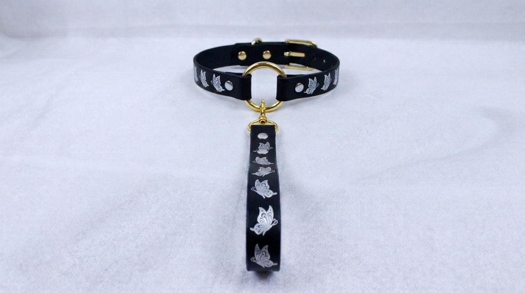 A black collar with a gold-colored buckle and a silver-colored tag featuring a repeating pattern of cats.