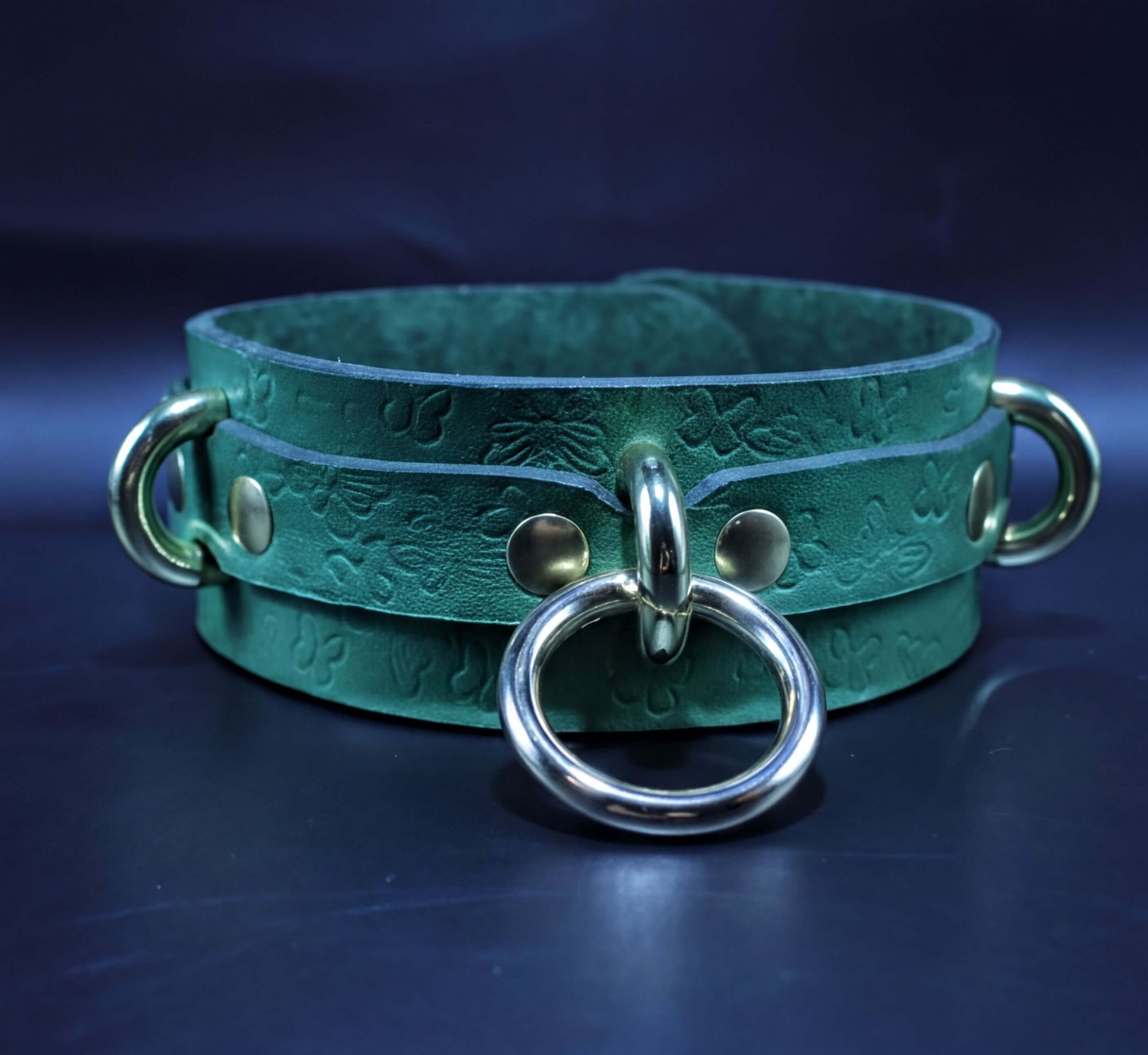 A green leather choker collar with metal rings and a buckle, displayed against a dark background.