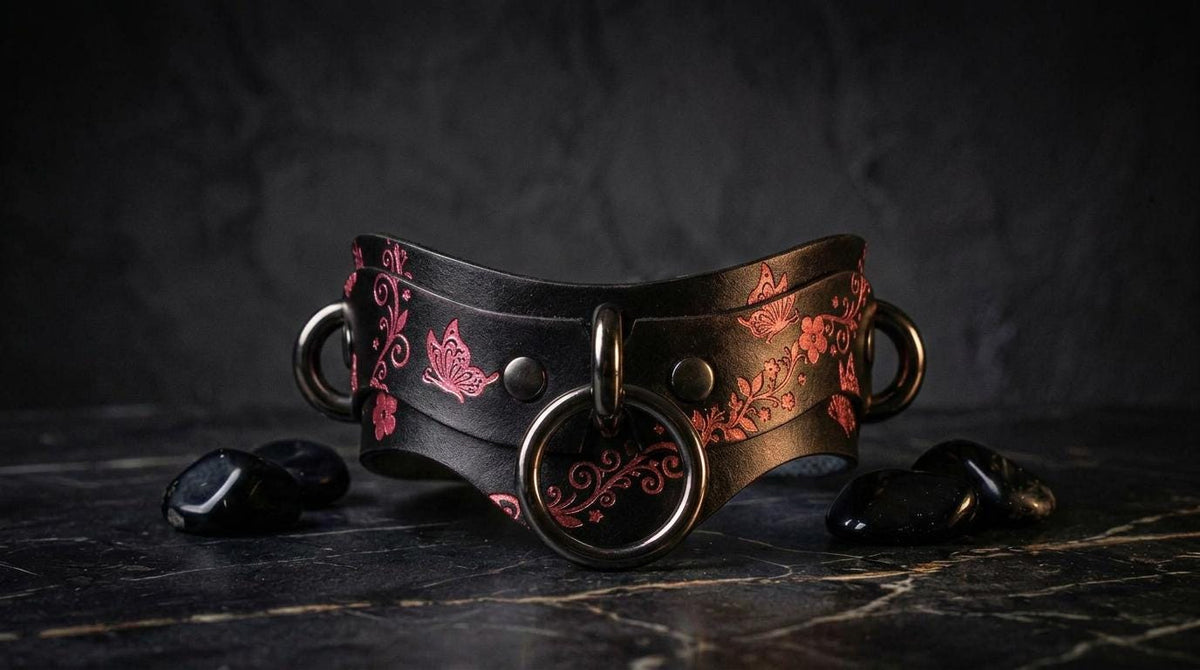 A black leather choker necklace with a floral pattern and metal rings is displayed on a dark surface, accompanied by black stones.
