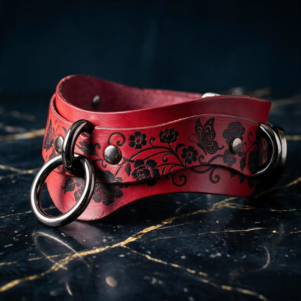 A red leather strap with black floral designs and metal rings is displayed on a dark surface.