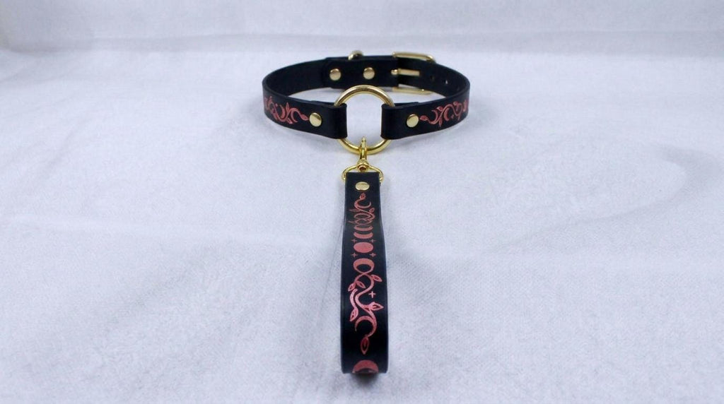A black leather collar with a gold-colored buckle and a decorative handle attached to it, resting on a white surface.