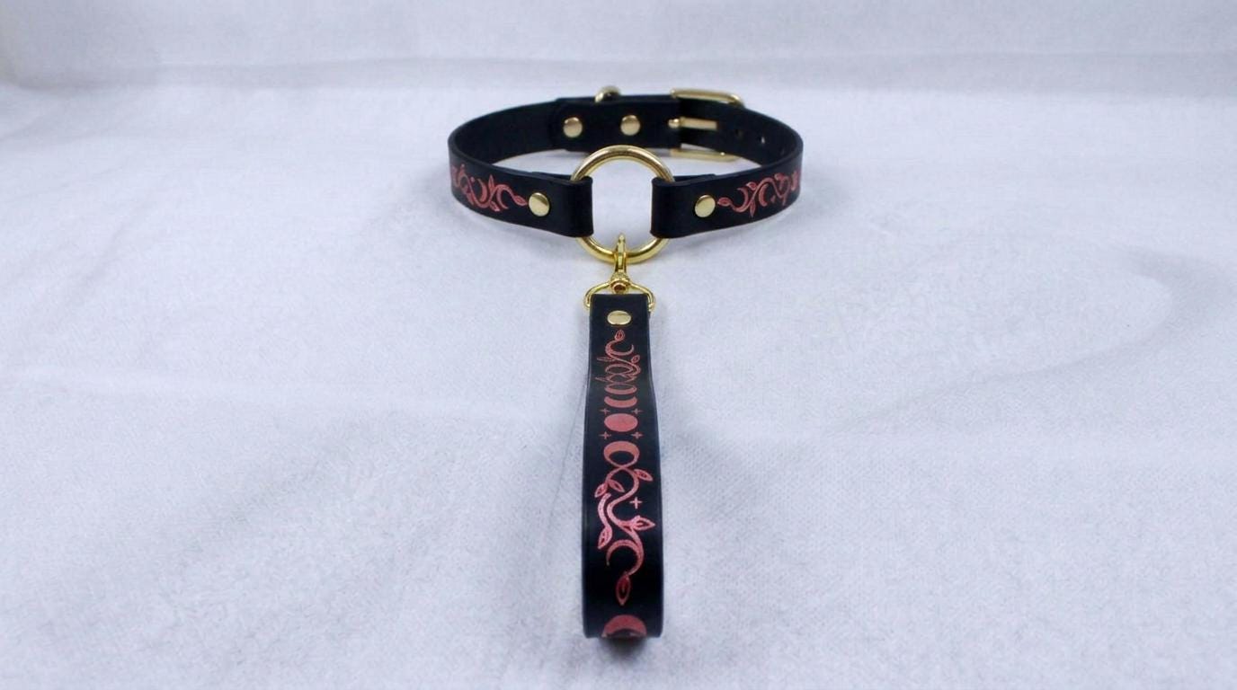 A black leather collar with a gold-colored buckle and a decorative handle attached to it, resting on a white surface.