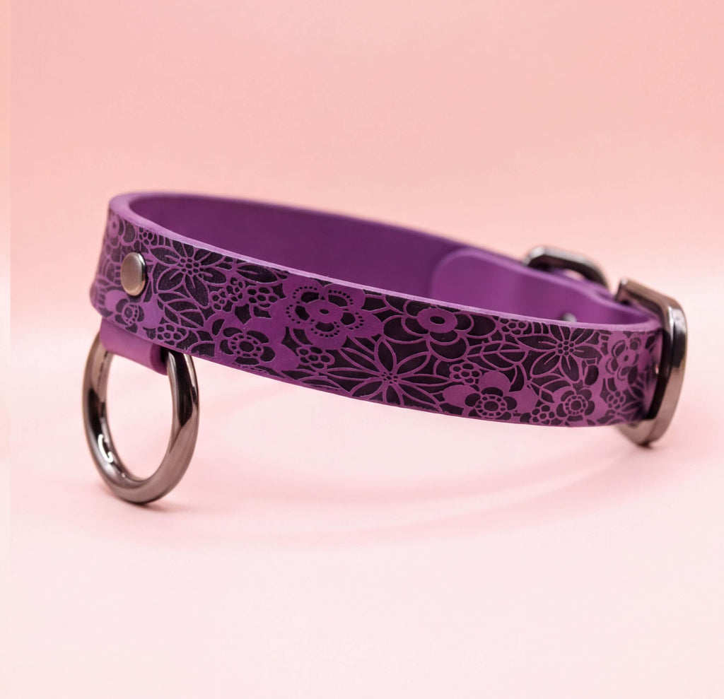 A purple dog collar with a floral pattern and metal rings on the sides.