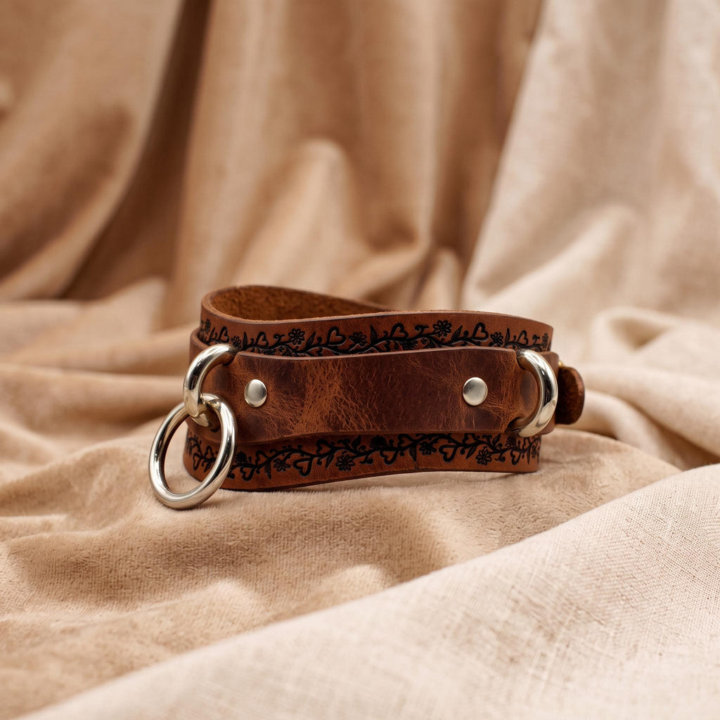 A brown leather choker necklace with metal rings and a decorative pattern on the clasp is displayed on a beige fabric background.