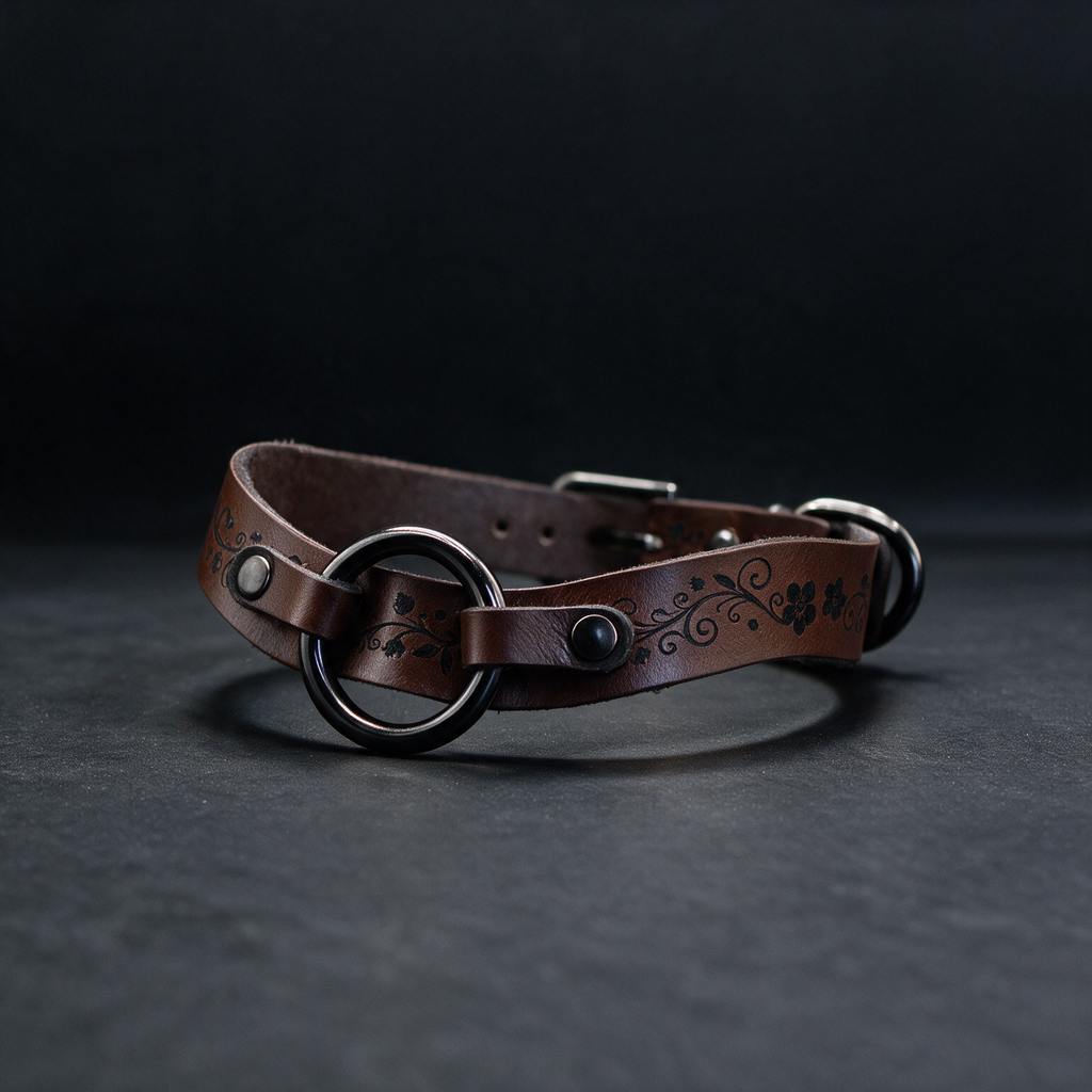 A brown leather collar with a decorative metal ring and floral pattern on it, resting on a dark surface.