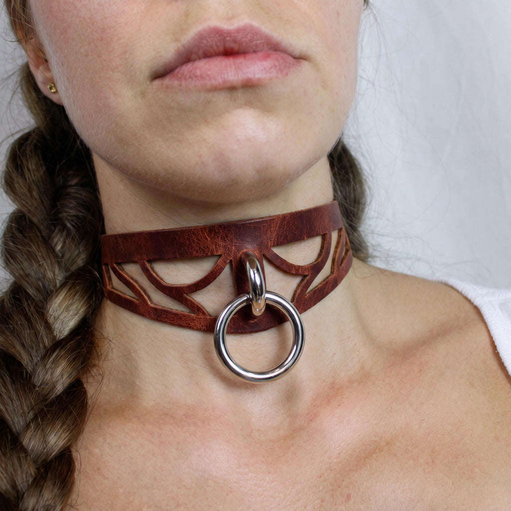 A close-up of a woman's neck and chest area, featuring a leather choker necklace with a metal ring and a braided hairstyle.