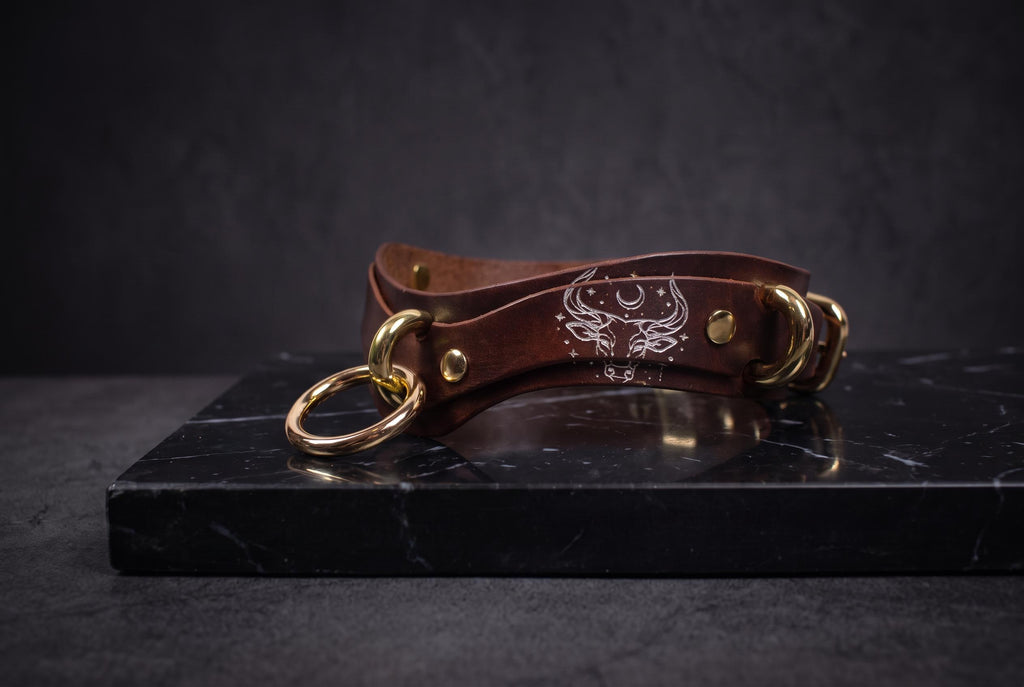 A brown leather collar with gold rings and a white design on the front, resting on a black marble surface.