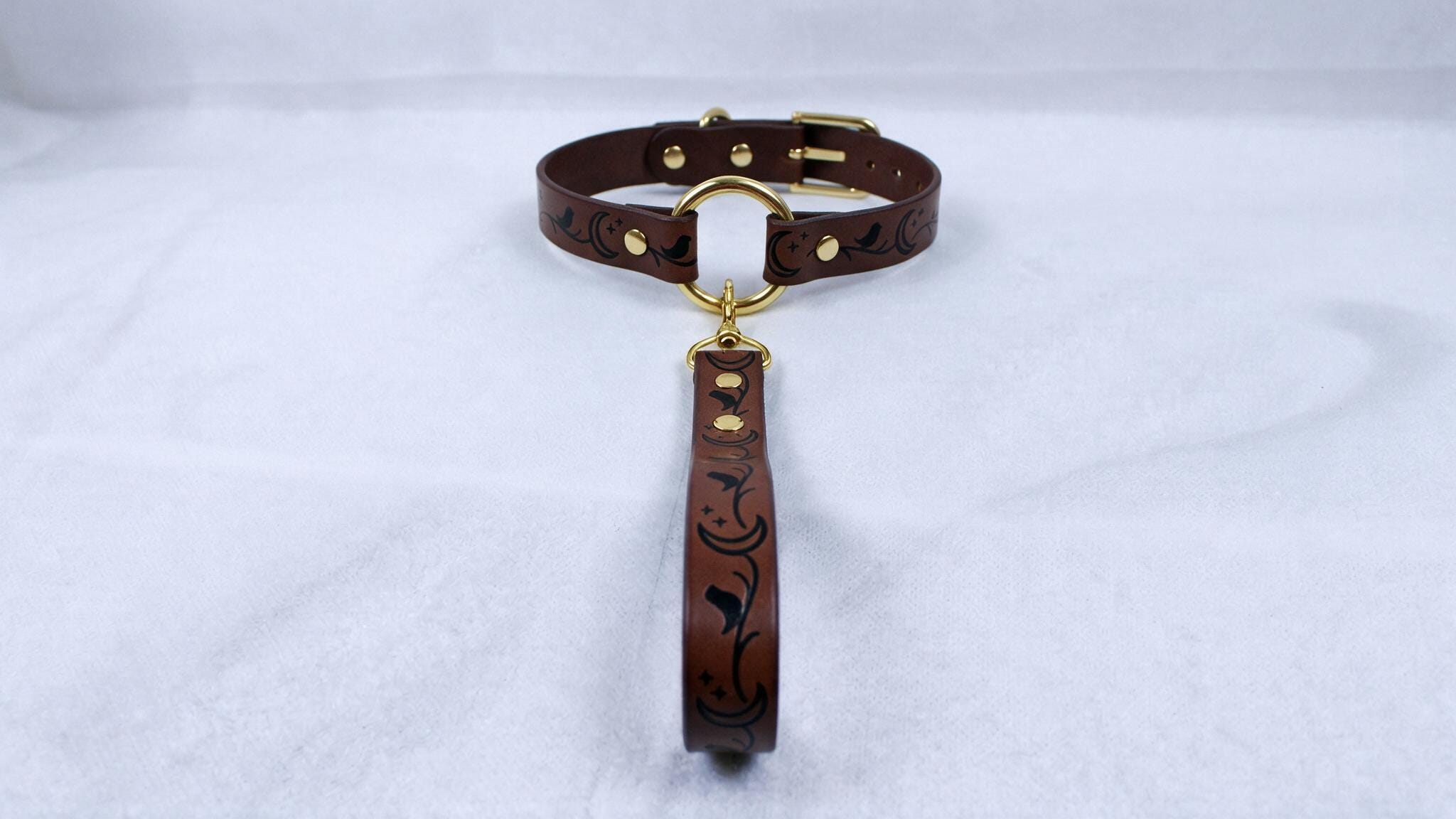 A brown leather collar with a gold-colored buckle and a decorative handle attached to it, resting on a white surface.