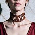 A close-up of a woman's neck and chest area wearing a large, leather choker necklace with the word "SLAVE" carved into it.