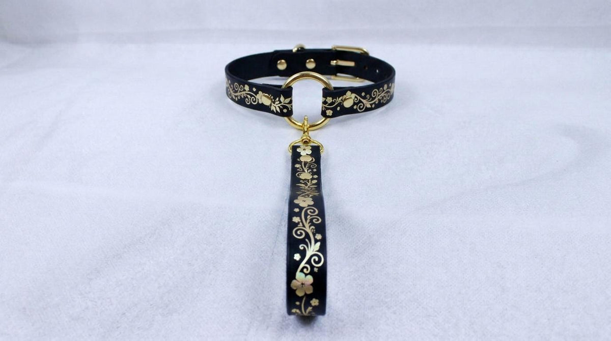 A black collar with gold floral designs and a gold-colored loop hanging from it.